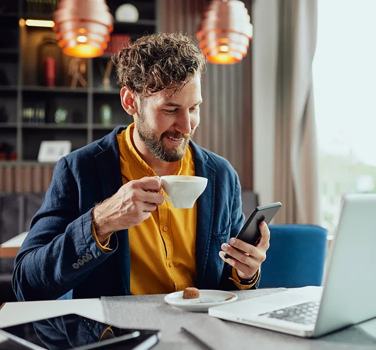 A man with a coffee checking his phone beside a laptop